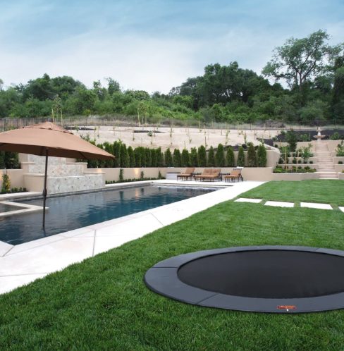 Round black in-ground trampoline set flush in a green lawn beside a swimming pool, with a patio umbrella and terraced landscaping beyond