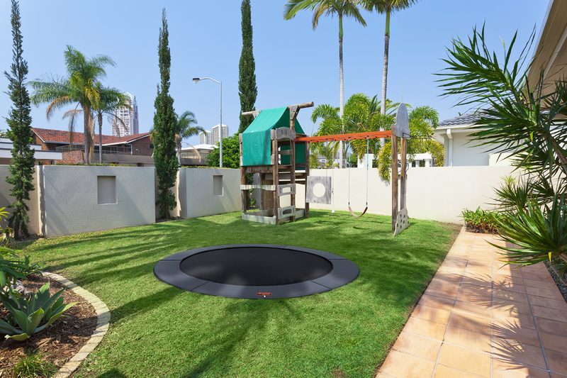 Round in-ground trampoline in a walled backyard lawn with a wooden play set, a paver path, and palm trees