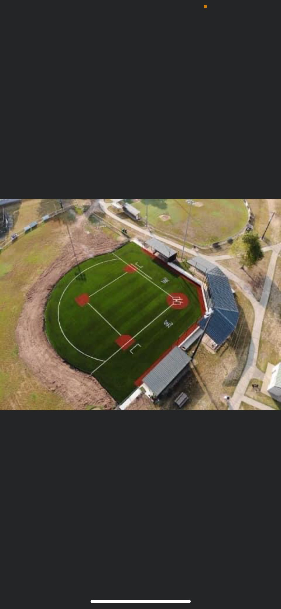 Aerial view of an artificial turf baseball or softball diamond with dugouts, a blue-roof backstop, white field lines, and a brown dirt-colored turf border
